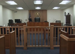 [ai] A view of an empty courtroom featuring wooden benches, a judge's bench with an ornate black chair, and a U.S. flag. A partition separates the seating area from the judge's space.